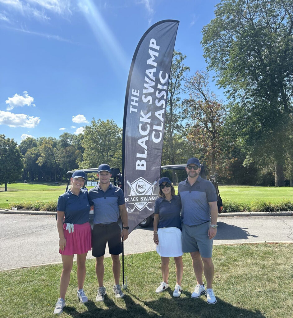Quatre personnes sur le parcours de golf du tournoi « Black Swamp Classic », à côté de la signalétique de la marque.